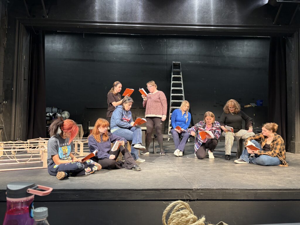 Nine women appear on a prescenium stage. Some are sitting, with two locked in a standing argument. Everyone has a script in hand. 