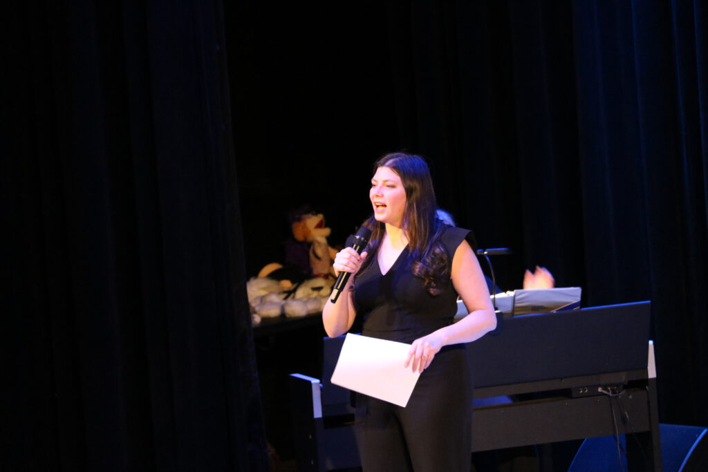 A woman in black excitedly reads from a list. She is holding a microphone.