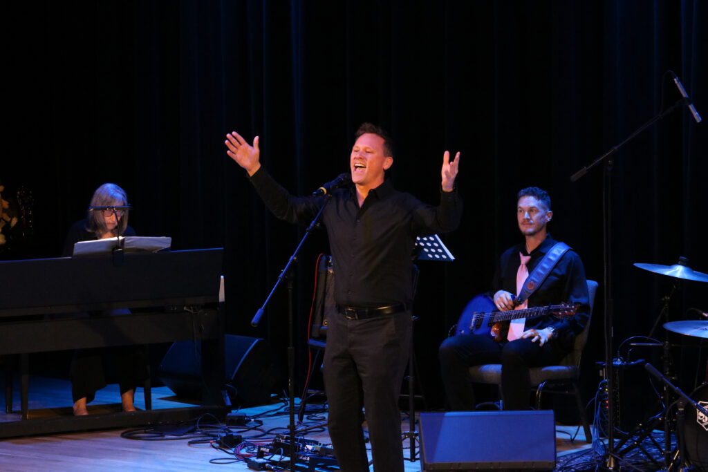 A man stands on stage singing into a microphone with arms outstretched. Behind him is a live band consisting of a guitarist and a pianist. All are wearing black.