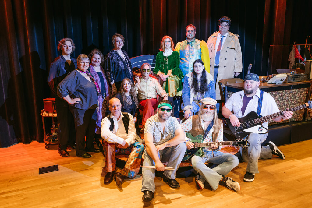 The cast and band of Fleetwood Macbeth smile for a picture. Some are wearing 70s inspired attire, whereas members of the tech crew wear blacks. They are of varying ages, but all smile brightly.
